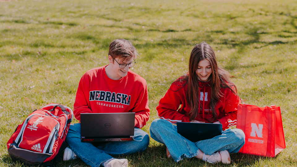 two people sitting on grass, smiling and typing on laptops