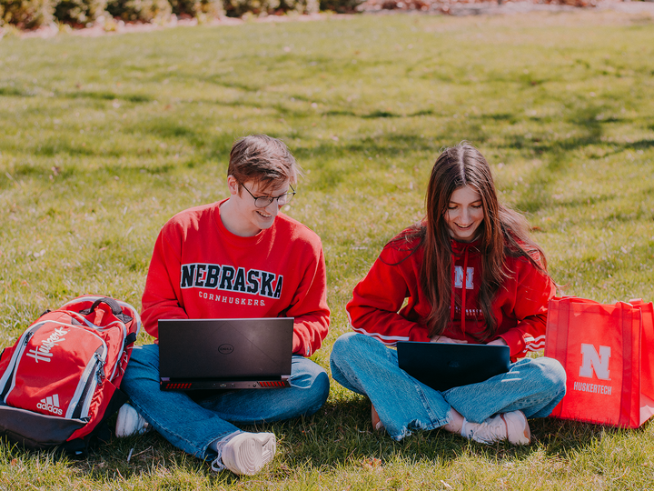 two people sitting on grass, smiling and typing on laptops